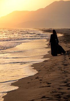 Silhouette of a woman walking on a serene beach during sunset with gentle waves.