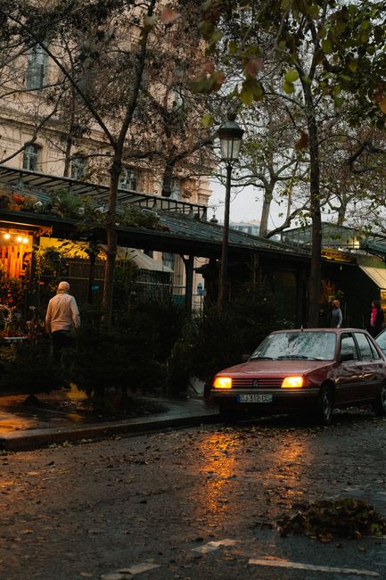 An urban street scene with a red car and people under dim streetlights, reflecting in rain-soaked pavement.