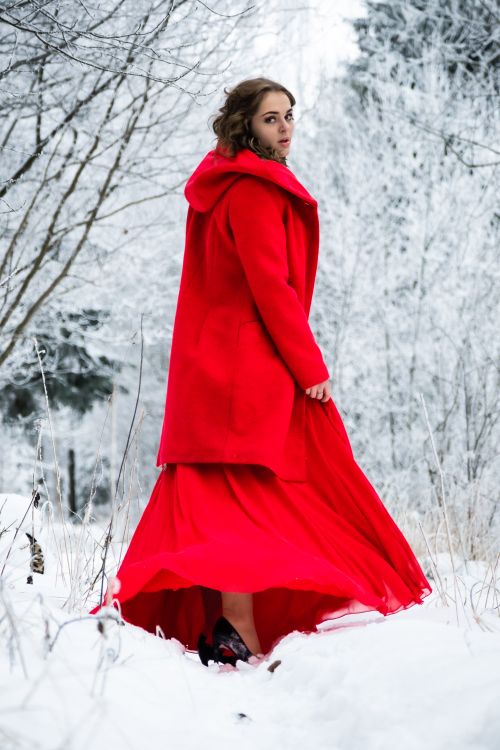 Free A woman in a striking red coat and dress walks through a snow-covered forest. Stock Photo