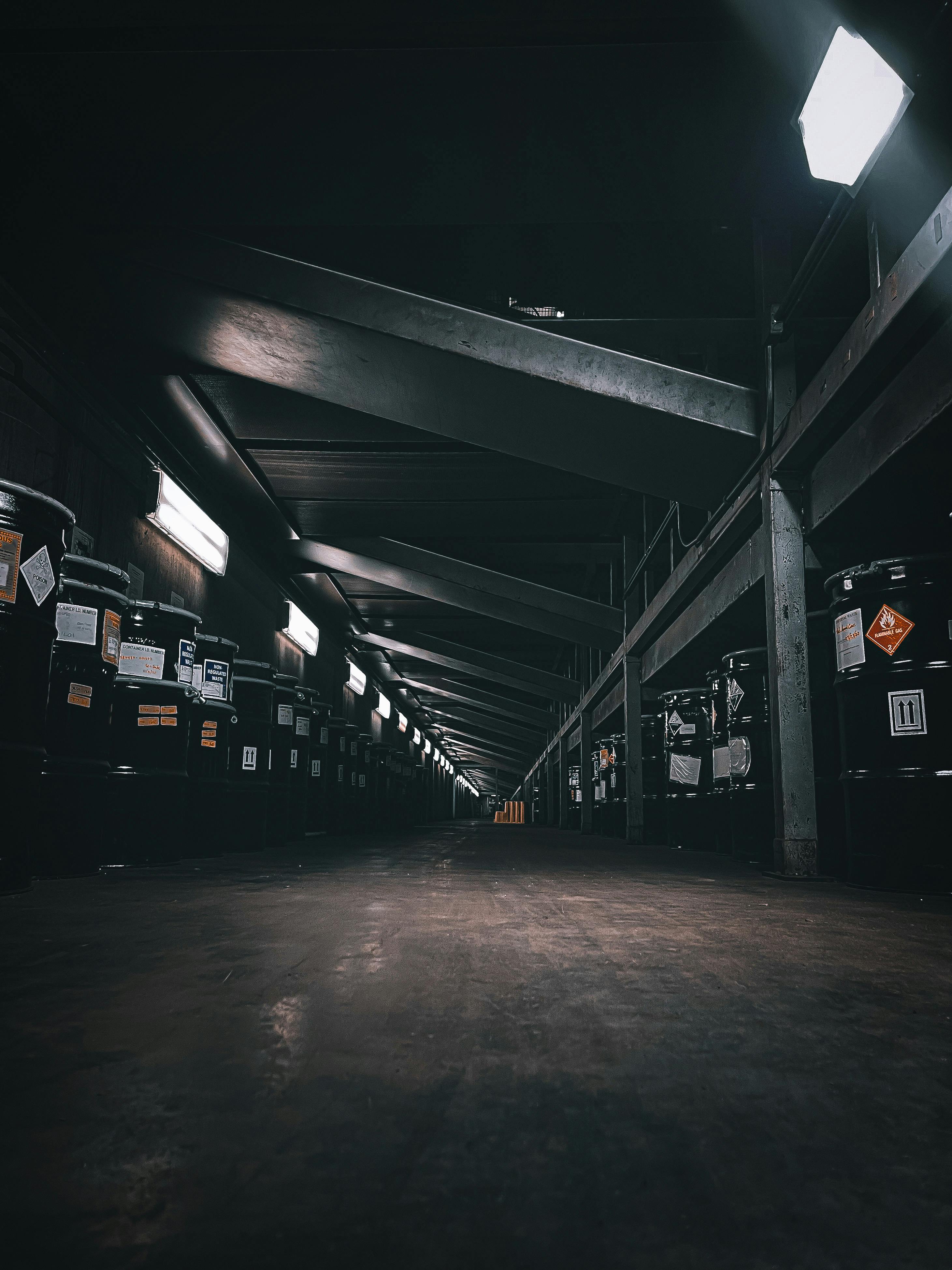 A dark, illuminated warehouse corridor with rows of storage barrels and beams overhead.