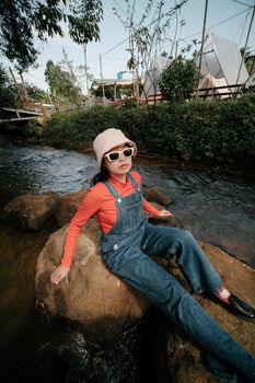 Stylish woman in bucket hat sitting by the riverside, showcasing casual fashion.