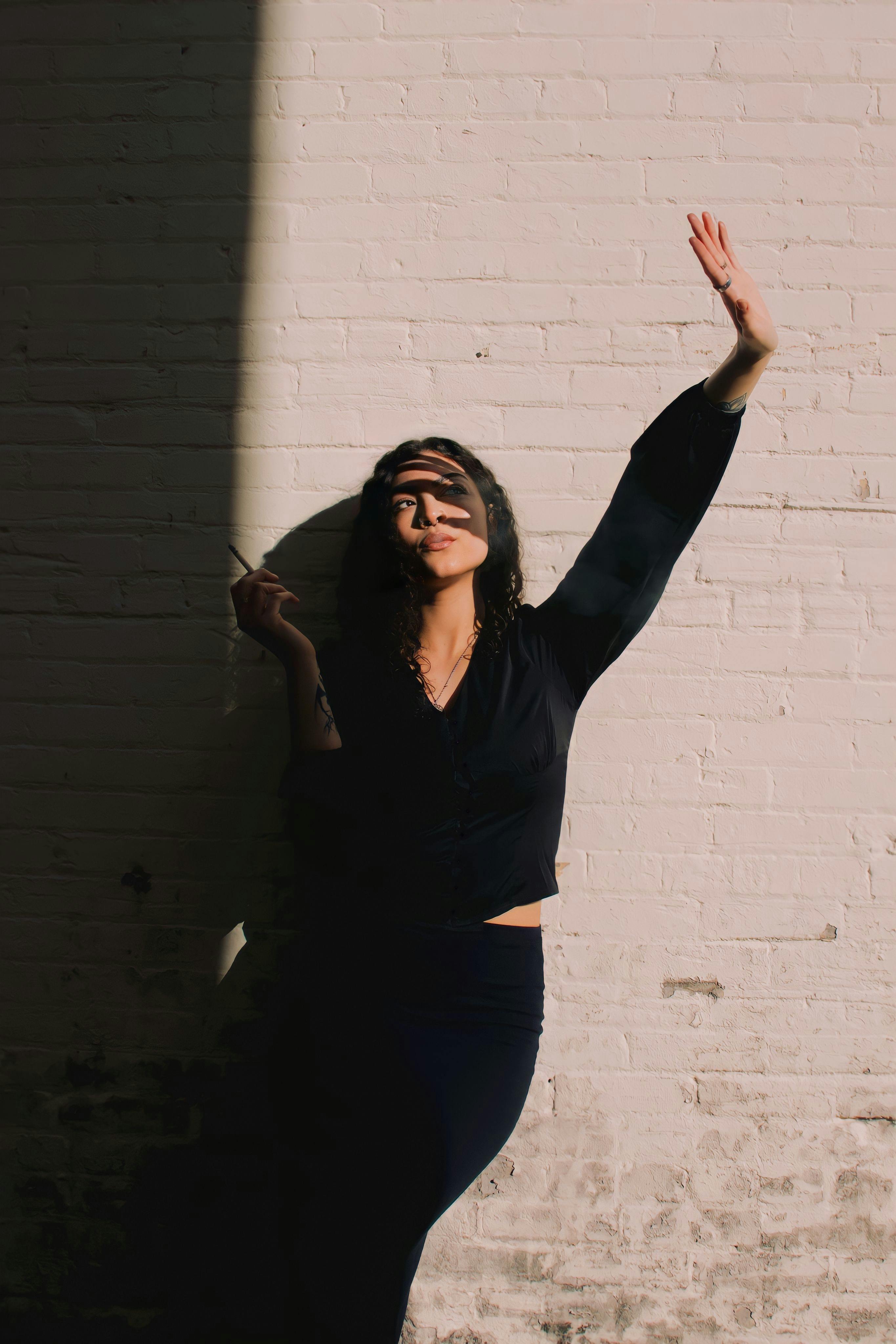 Woman in black clothing poses against a brick wall with striking shadows.