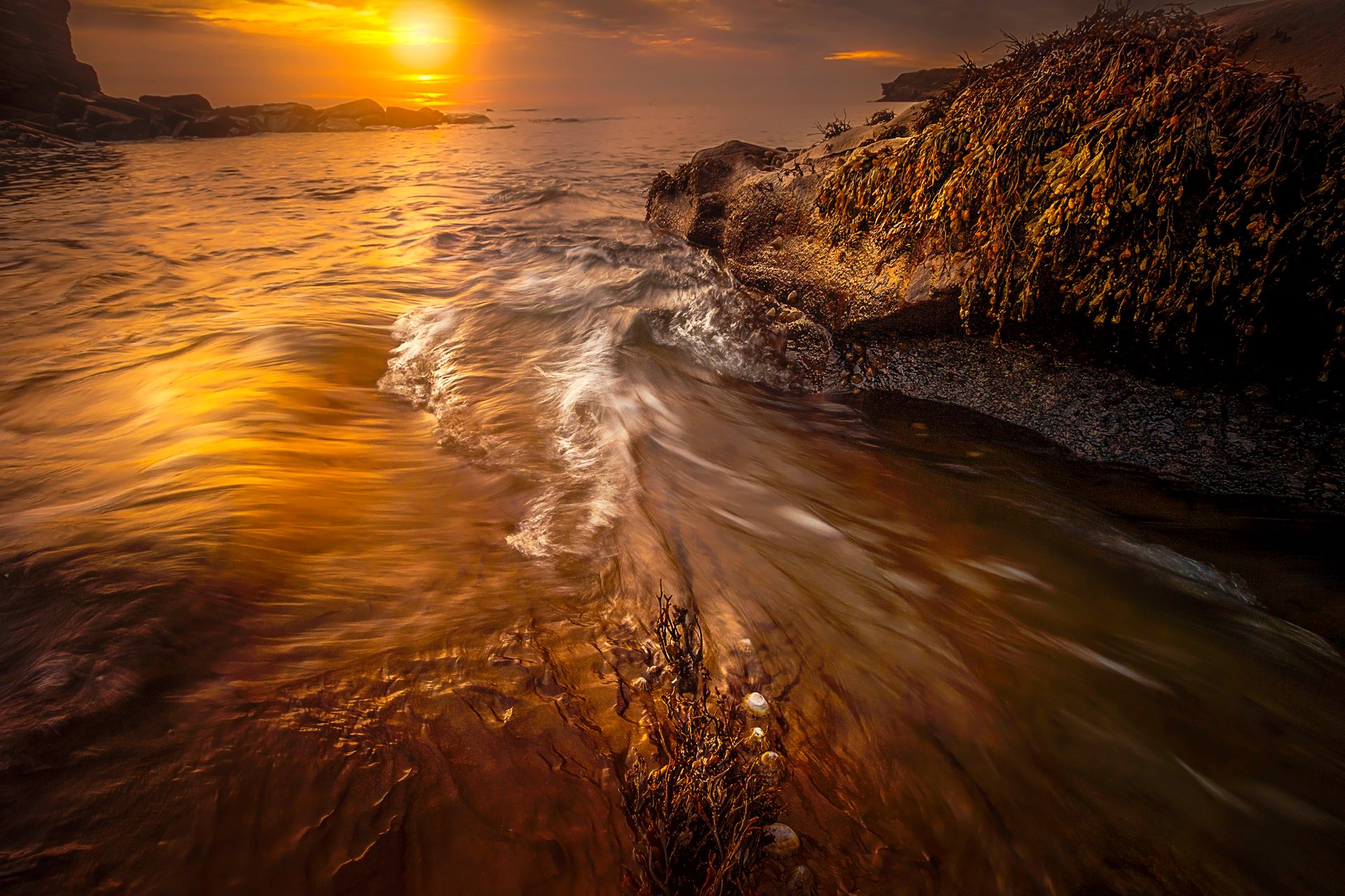 Sunset view of ocean waves flowing over rocky shore with golden reflections.
