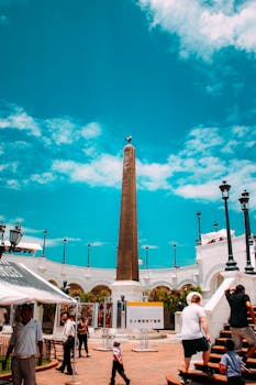 Tourists explore a sunny plaza in Panama City featuring a tall obelisk and historical architecture.