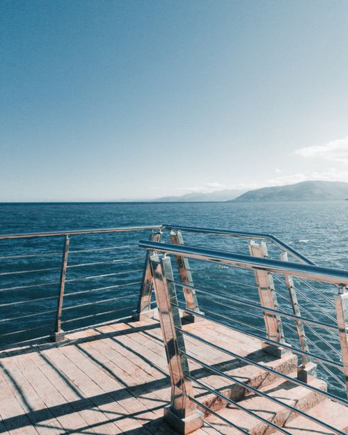 Free Scenic view of a boardwalk with railings, overlooking the vast blue sea with distant mountains. Stock Photo