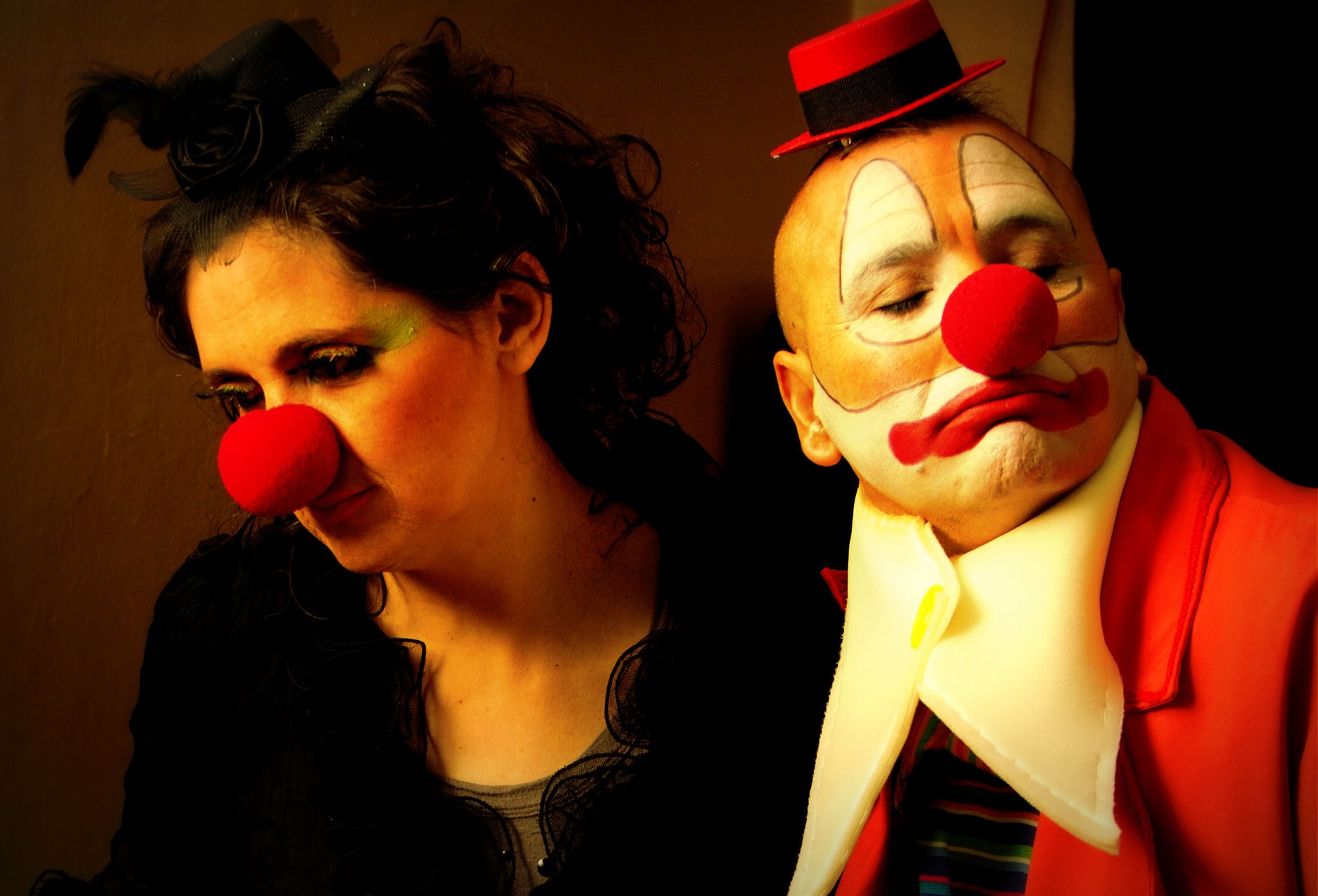Two clowns with dramatic makeup and red noses pose with solemn expressions against a neutral backdrop.