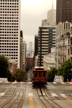 Historic cable car on a steep San Francisco street, cityscape with skyscrapers and the Bay Bridge in the background, urban transportation.