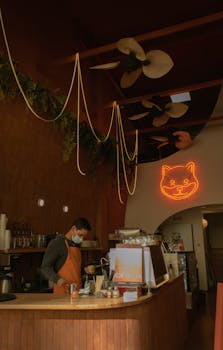 A barista prepares coffee in a warm, inviting café with a cat-themed neon sign.