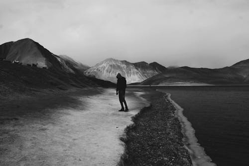 Grayscale Photo of a Person Standing on the Sea Shore near Mountains