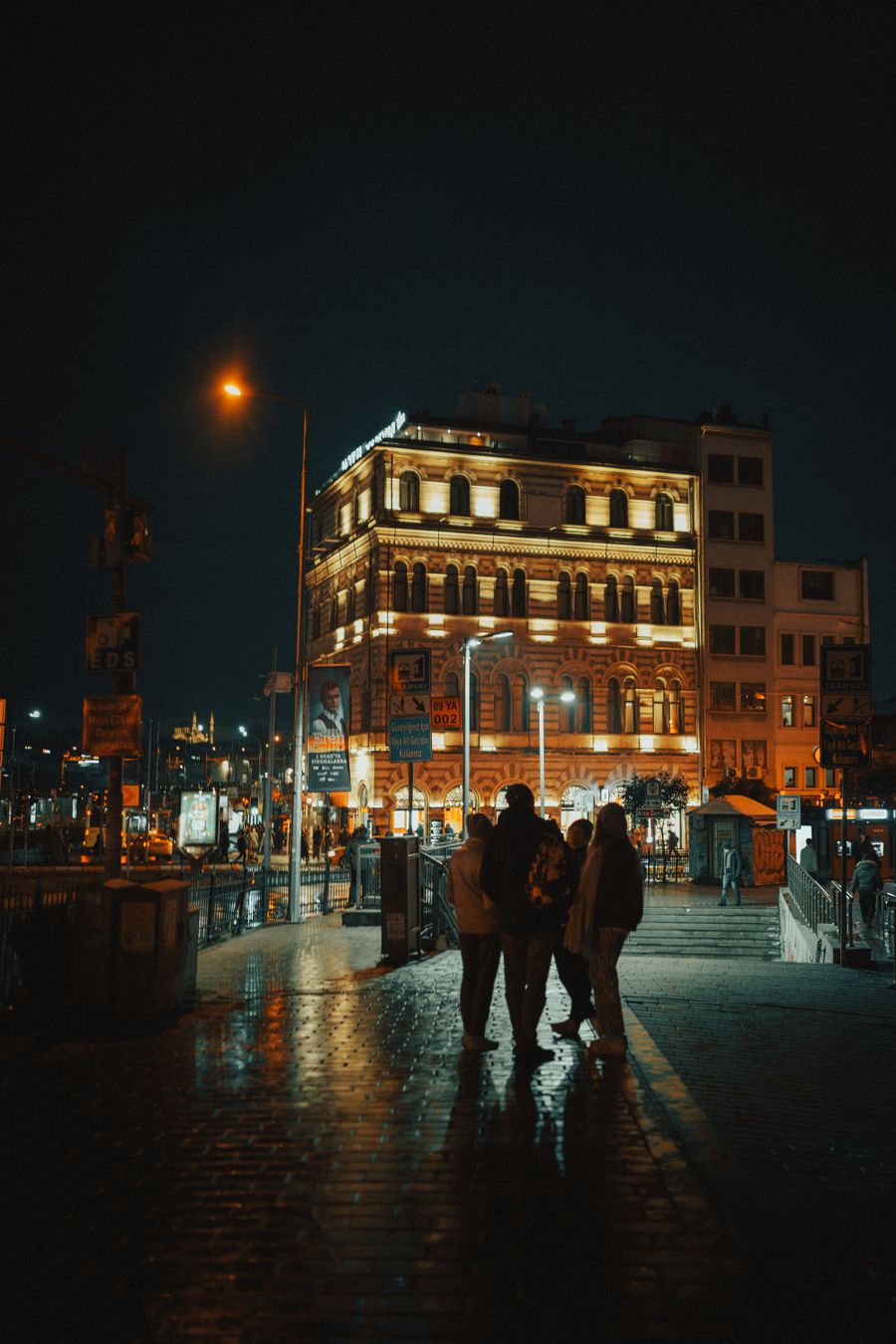 Gece ıslak bir sokakta ışıklı bir bina ve yayalar