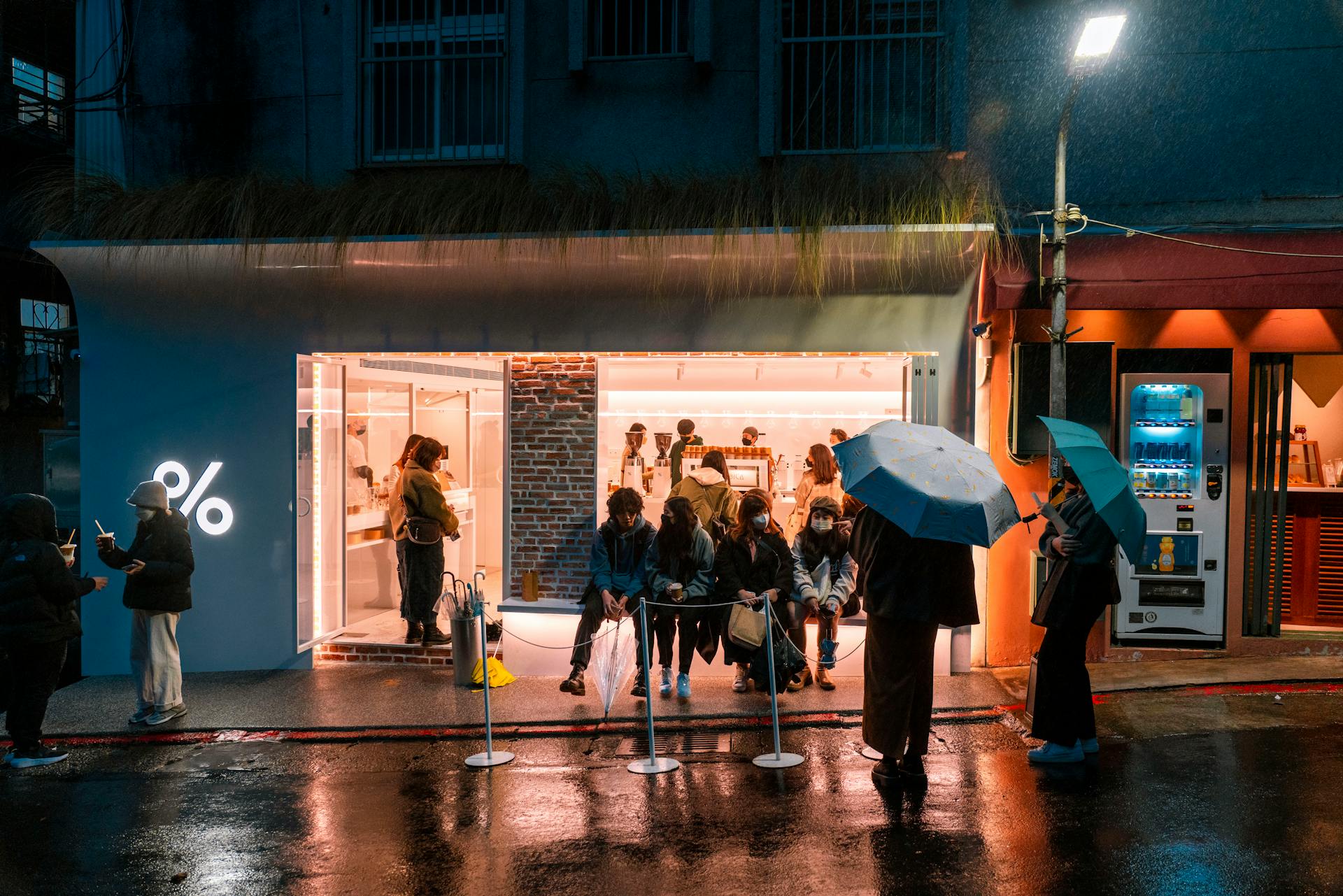 City street scene with people waiting outside a shop in the rain, illuminated signs and reflections.