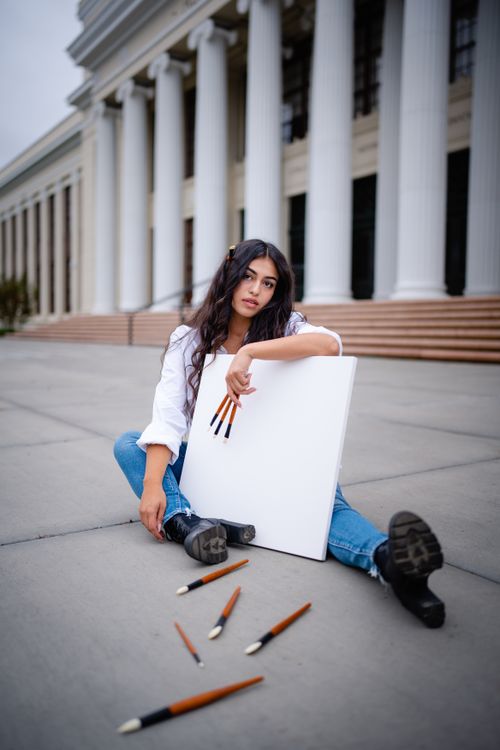 Free A young woman with brushes poses outdoors in front of a historic building, expressing creative contemplation. Stock Photo