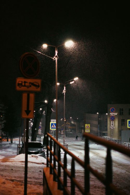 Free A serene winter night street scene with falling snow and illuminated street lamps in a quiet town. Stock Photo