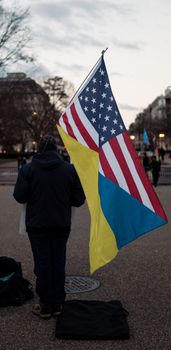 Person holds USA and Ukraine flags during twilight in an urban area.