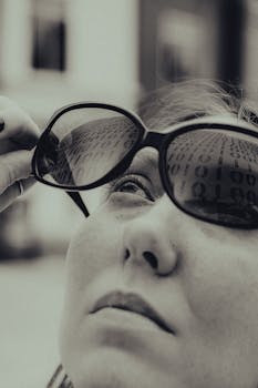 Black and white photo of a woman looking up with binary code reflected in sunglasses.