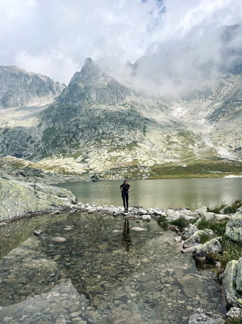 Person Standing near Lake in Mountains under Clouds