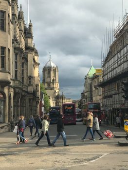 Pedestrians crossing a busy street in Oxford with historic architecture and iconic buses in view.