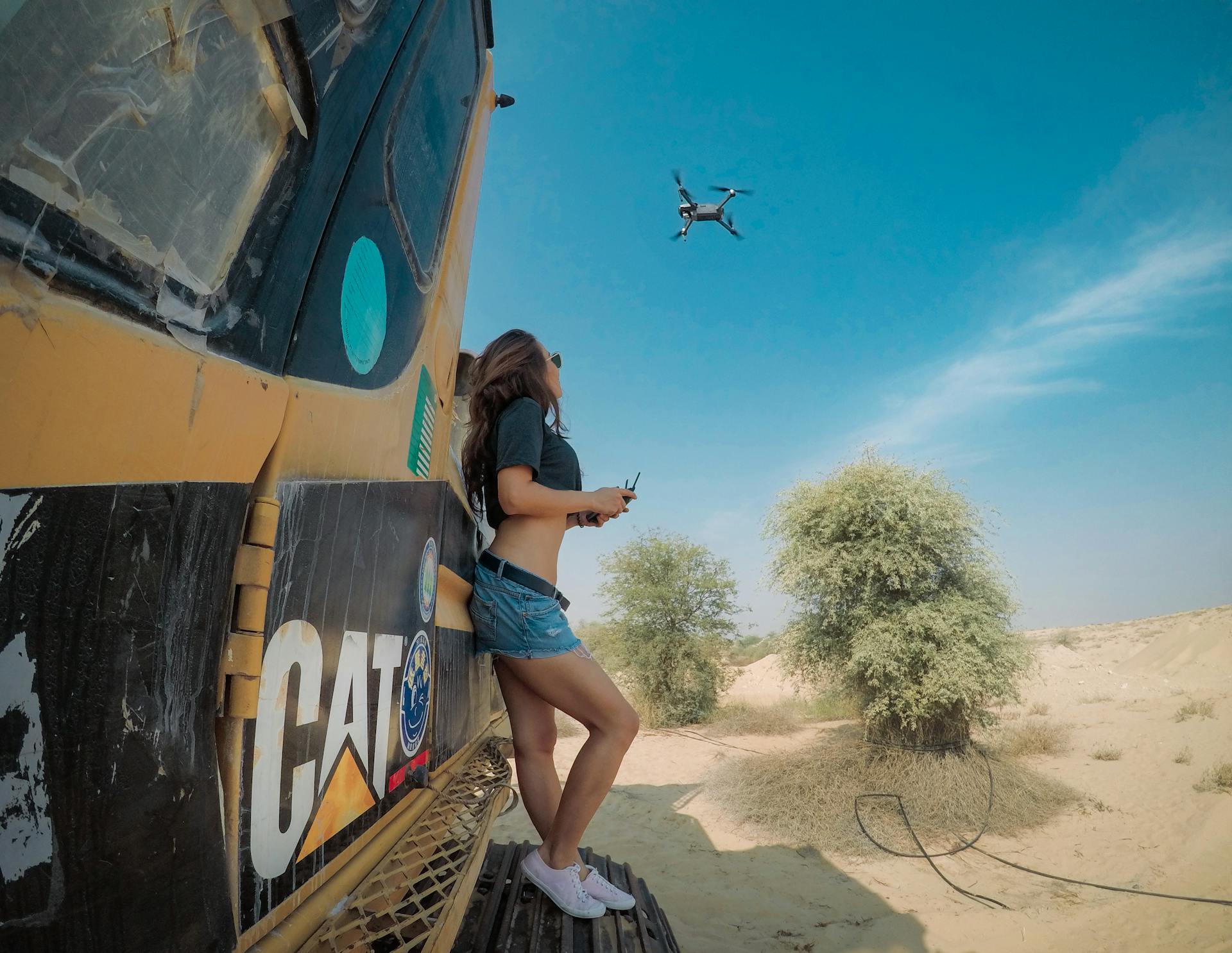 Woman flying drone above desert near excavator, Dubai skyline in view.