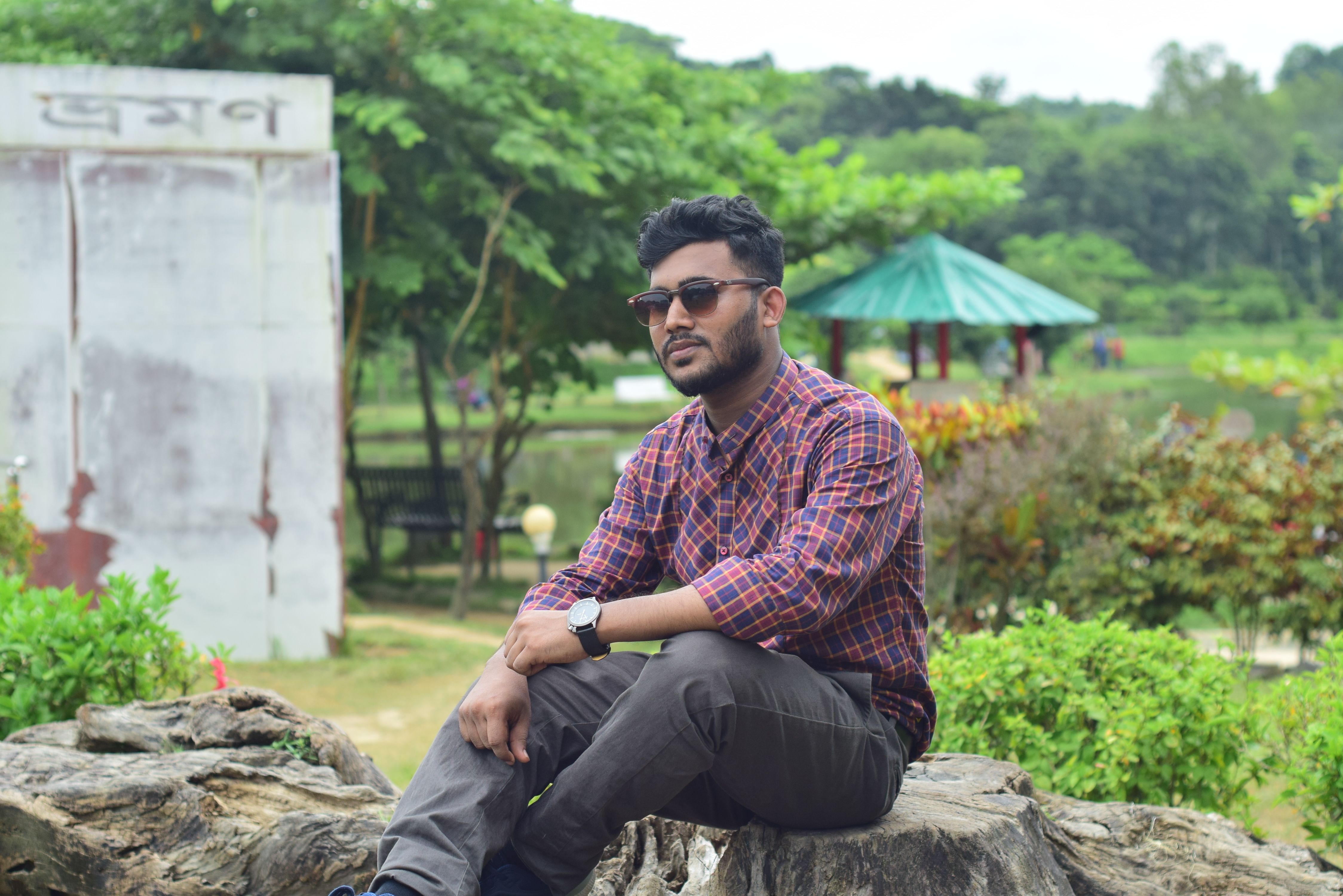 A stylish man wearing sunglasses sits outdoors on a rock with greenery in the background.