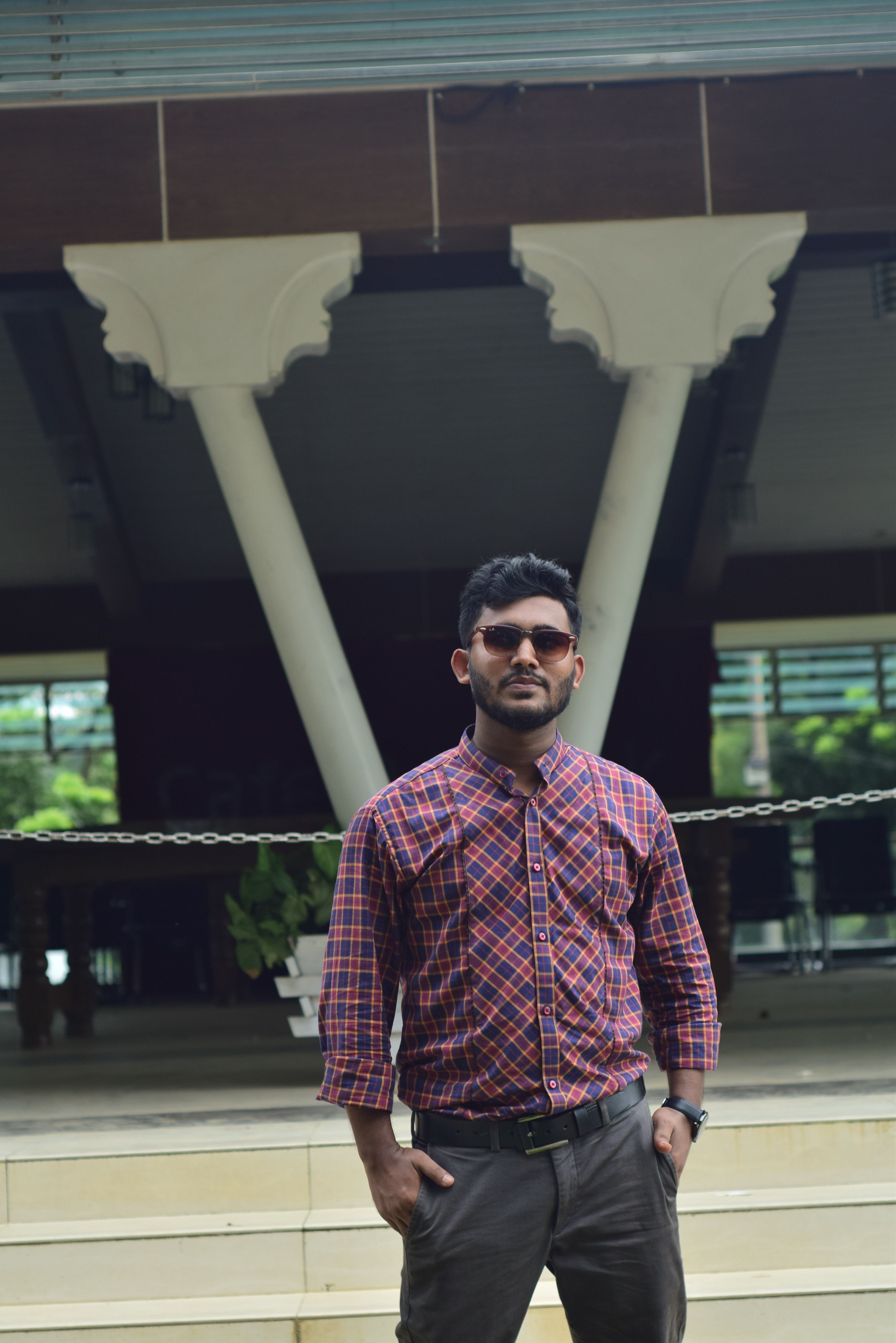 Confident man with sunglasses poses stylishly outdoors in a plaid shirt.