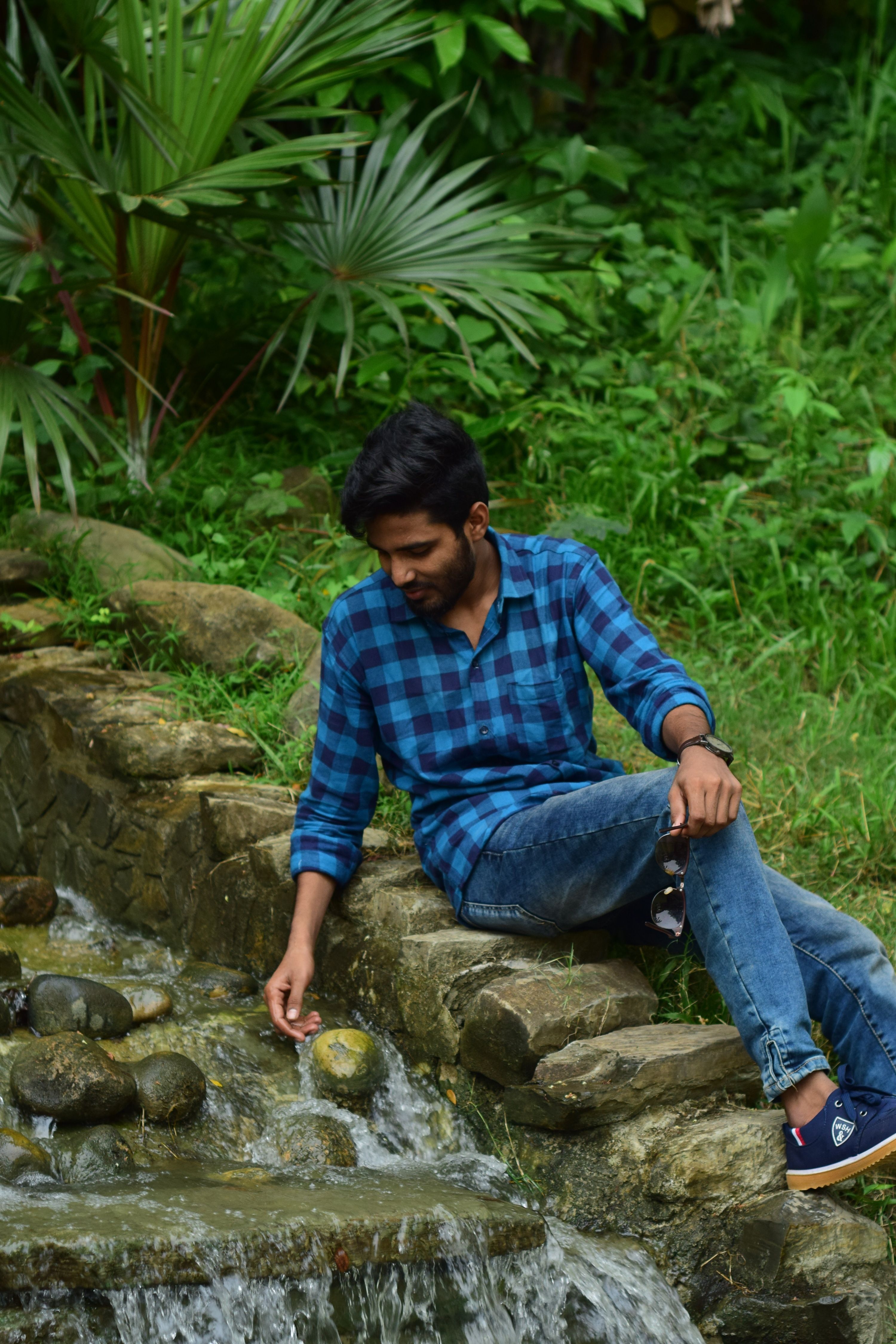 Asian man in a blue plaid shirt sitting by a creek, enjoying nature.