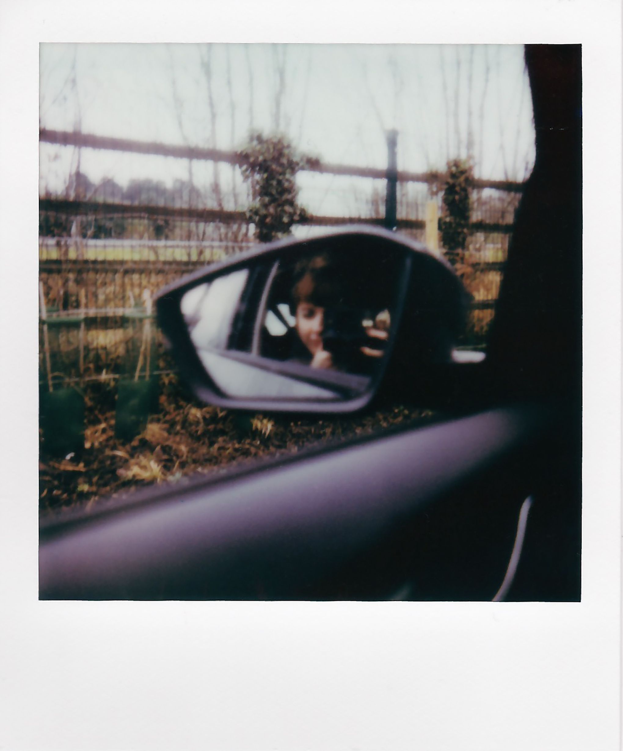 A reflection in a car mirror showing a person taking a photo with a fence in the background.
