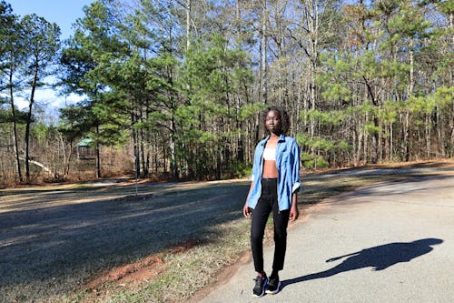 Woman in Blue Shirt, White Bandeau Top, and Black Jeans Walking on a Road