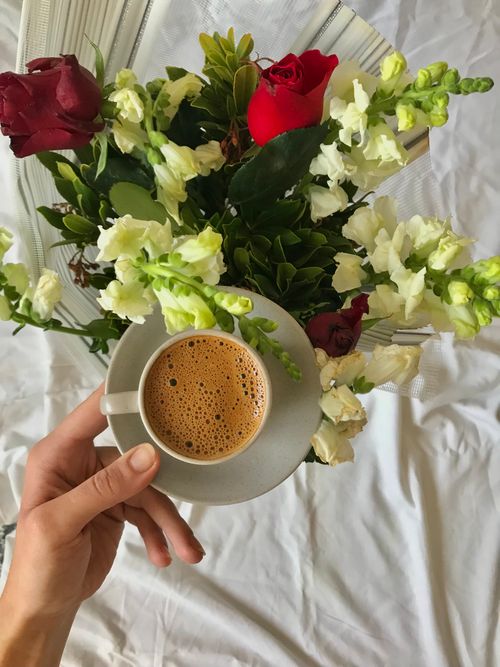 Free Close-up of a hand holding a coffee cup against a backdrop of red and white flowers arranged beautifully. Stock Photo