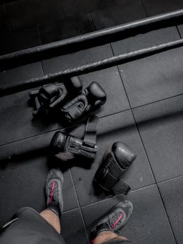 Close-up of black boxing gloves and athletic shoes on gym floor, signifying exercise and fitness.