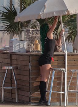 Woman adjusting a beach umbrella at an outdoor bar with tropical decor.