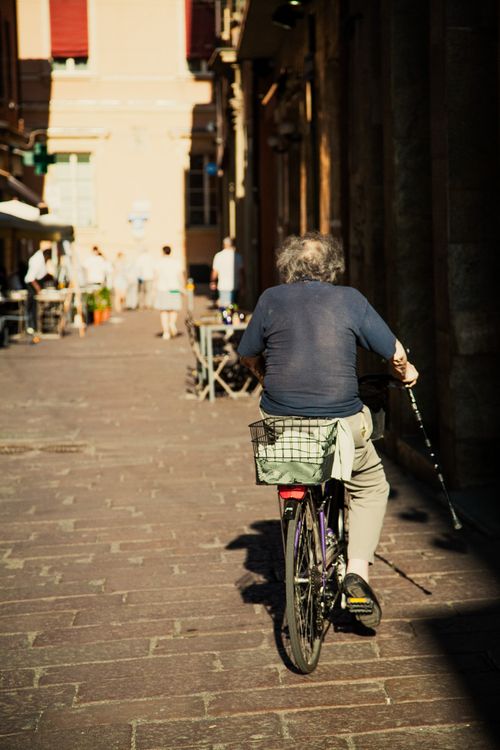 Free Senior adult cycling through a sunlit street in Bologna, Italy. Back view. Stock Photo