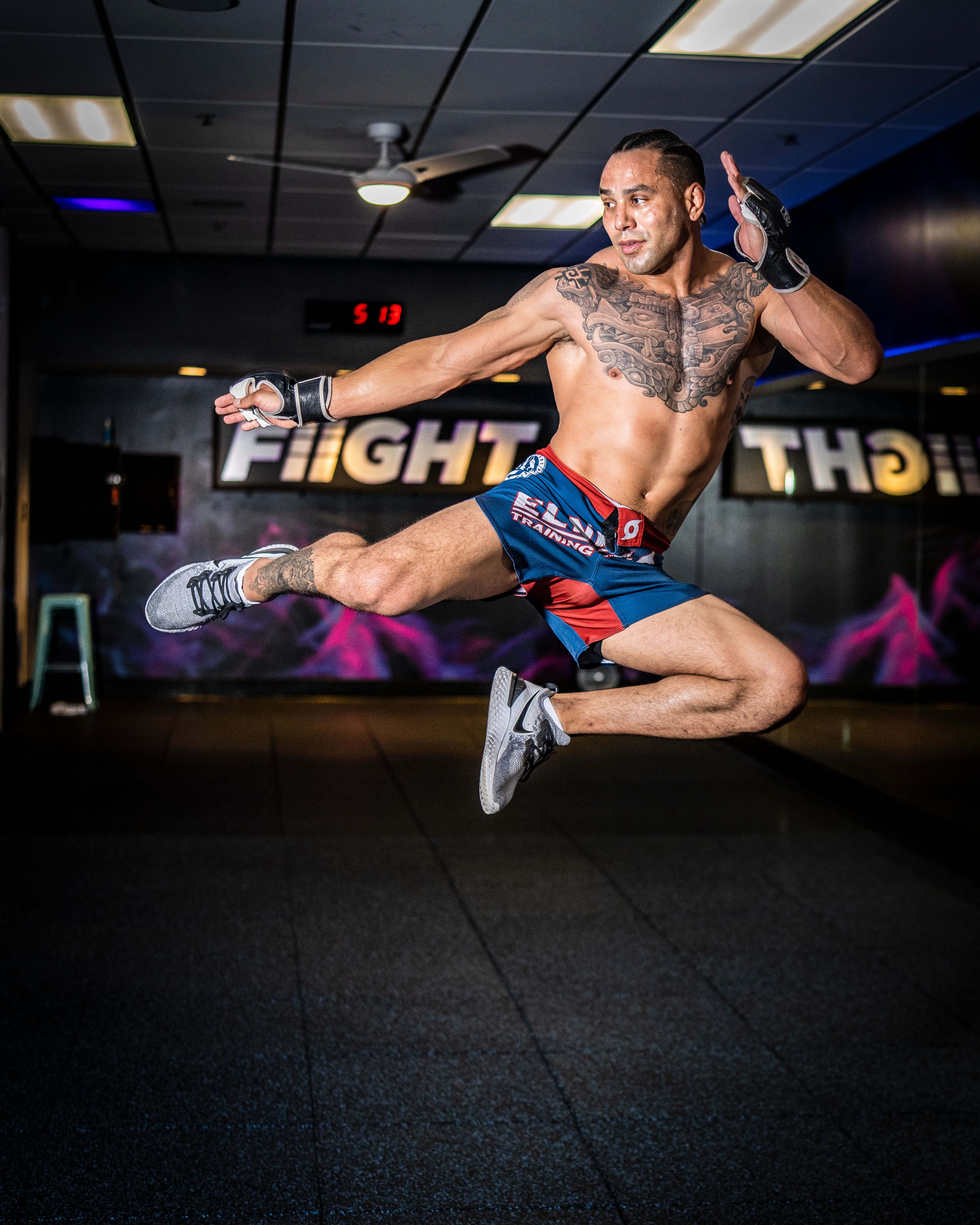 A tattooed man executes a high jump kick in a gym, showcasing athleticism and martial arts skill.