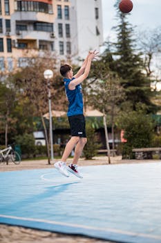 A teenager skillfully shoots a basketball outdoors on a vibrant blue court in an urban setting.