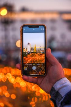 A person holds a smartphone capturing a vibrant city scene with decorative lights.