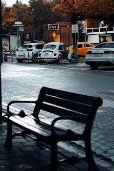 A vintage car and taxi sign on a quiet urban street with a wooden bench in the foreground.