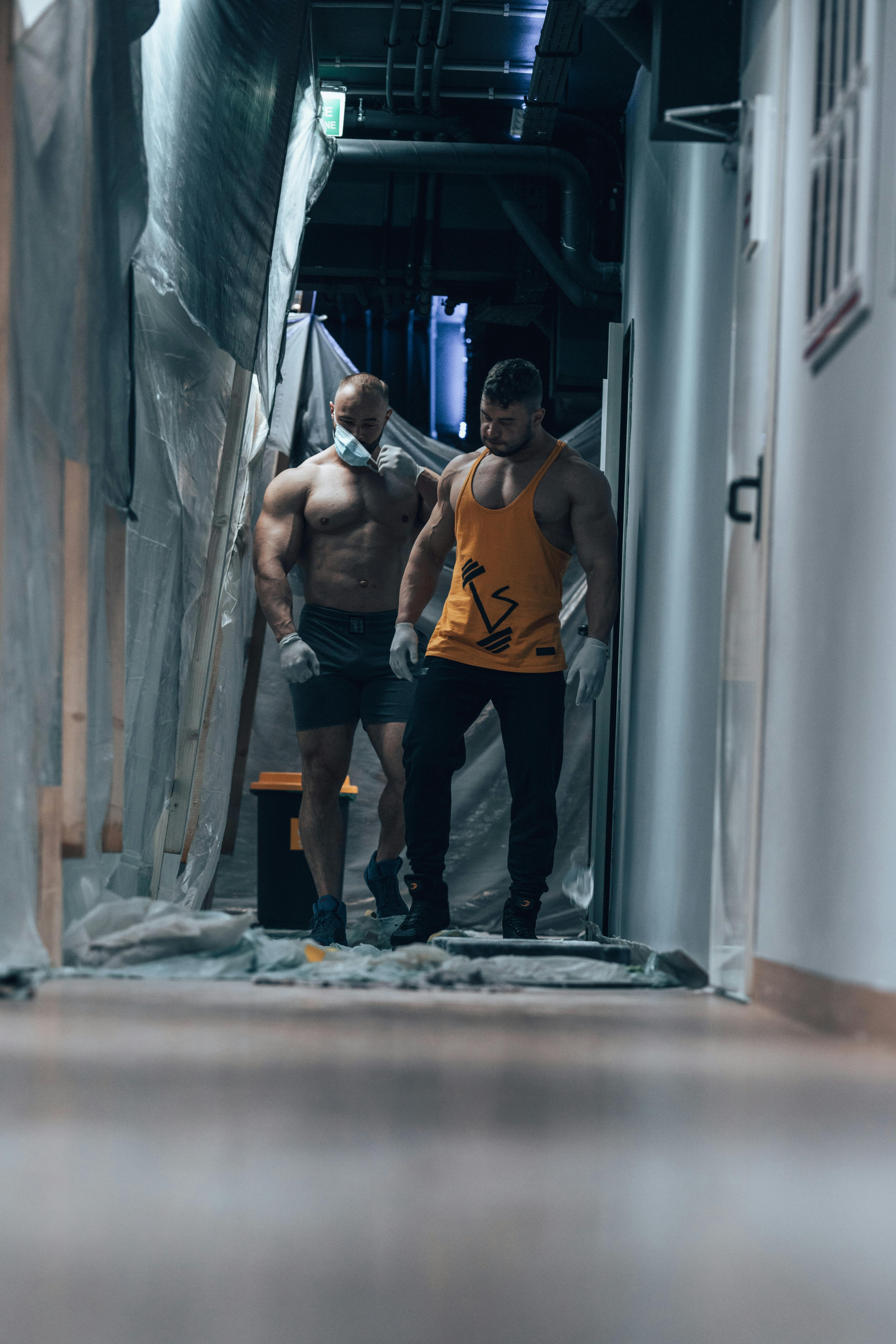 Two muscular men wearing gloves walk through a dimly lit corridor with plastic sheets.