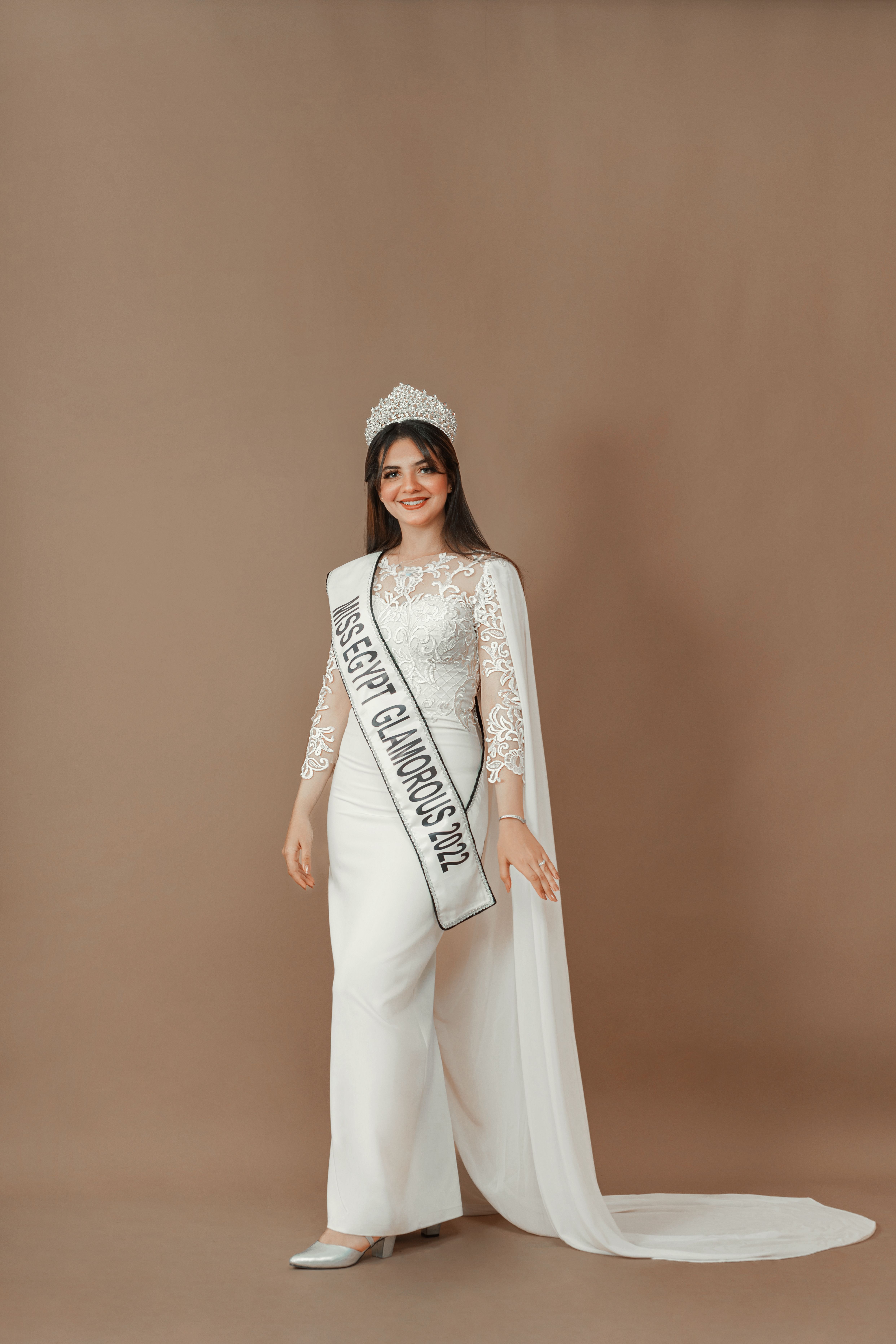 Elegant portrait of a beauty pageant winner in a white lace dress with a crown and sash, posing agai