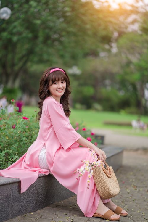 Free Woman in a flowing pink dress sitting on a bench holding a straw handbag in a lush park. Stock Photo