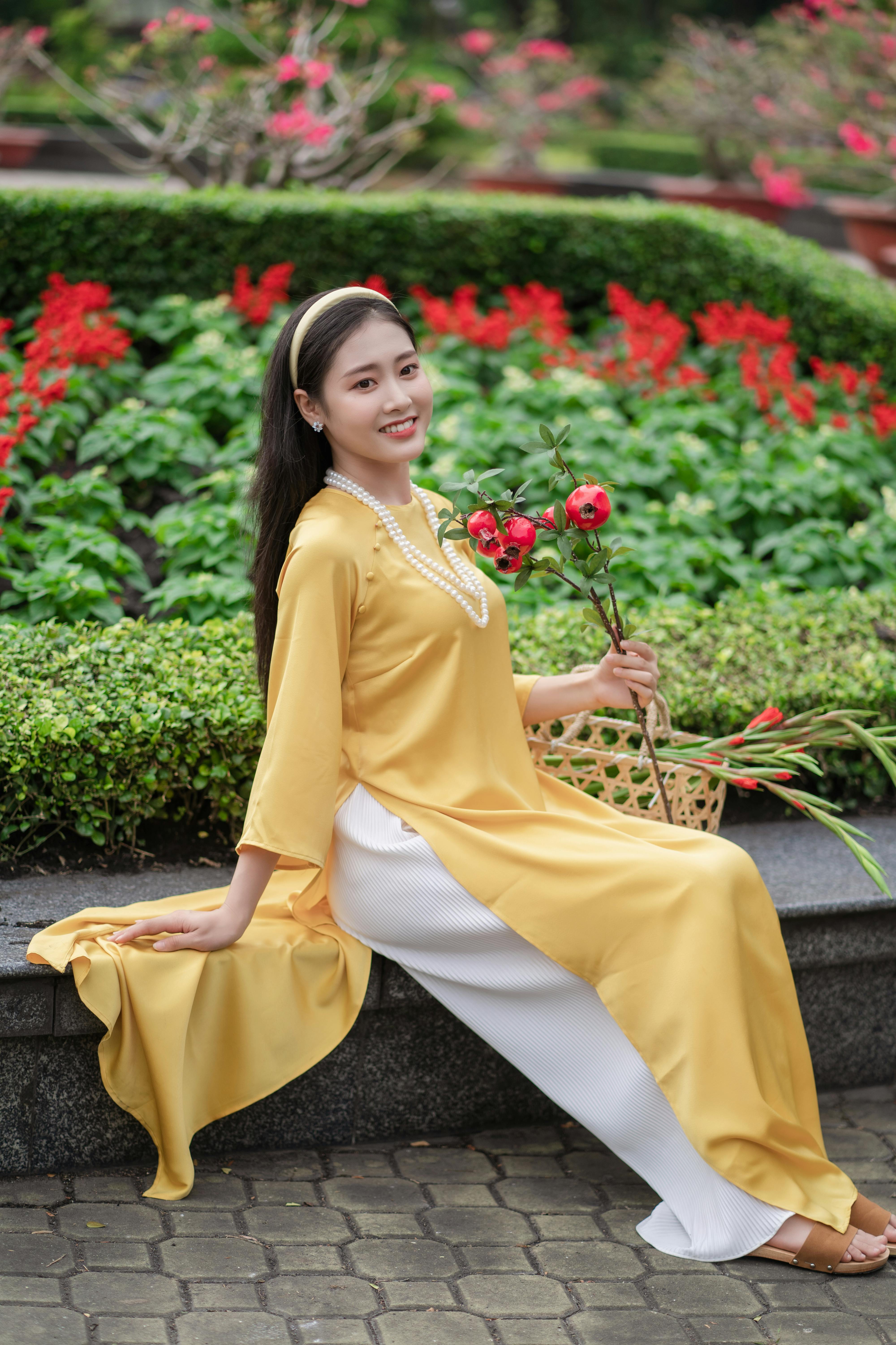 An Asian woman in a yellow ao dai smiles as she poses elegantly in a vibrant garden.