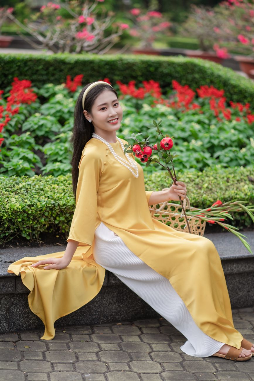An Asian woman in a yellow ao dai smiles as she poses elegantly in a vibrant garden.