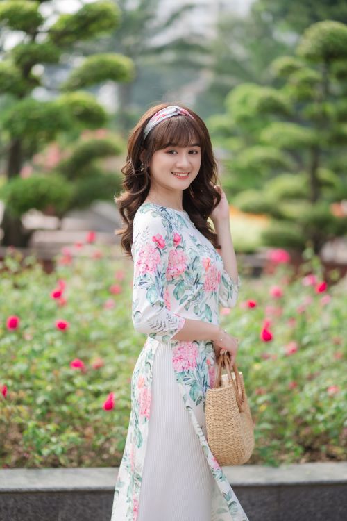 Free Asian woman in floral dress smiling in a park during summer, surrounded by flowers and trees. Stock Photo