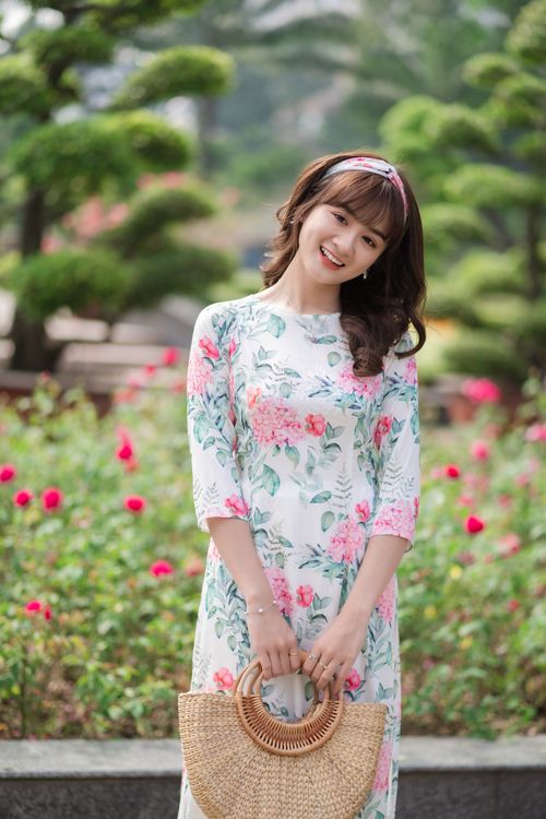 Free Young woman smiling in a floral dress holding a straw bag in a colorful garden. Stock Photo