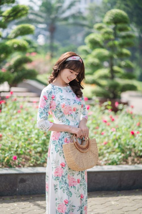 Free A young Asian woman in a floral ao dai poses gracefully with a straw bag in a vibrant garden. Stock Photo