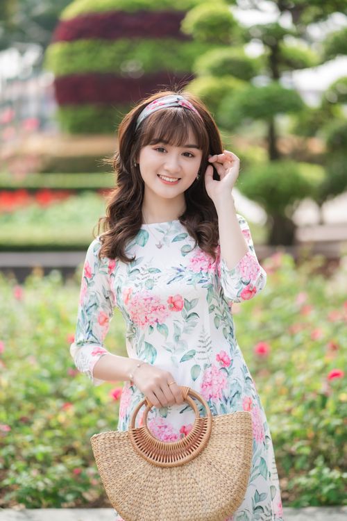 Free Smiling woman in a floral dress stands in a vibrant park, holding a straw handbag. Stock Photo