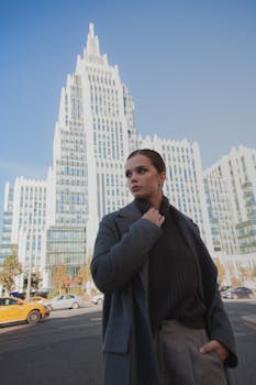 Stylish woman in warm clothing poses with a striking skyscraper in the background.