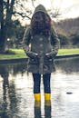 一名女子身穿绿色外套和黄色靴子，站在宁静的池塘中，四周环绕着大自然