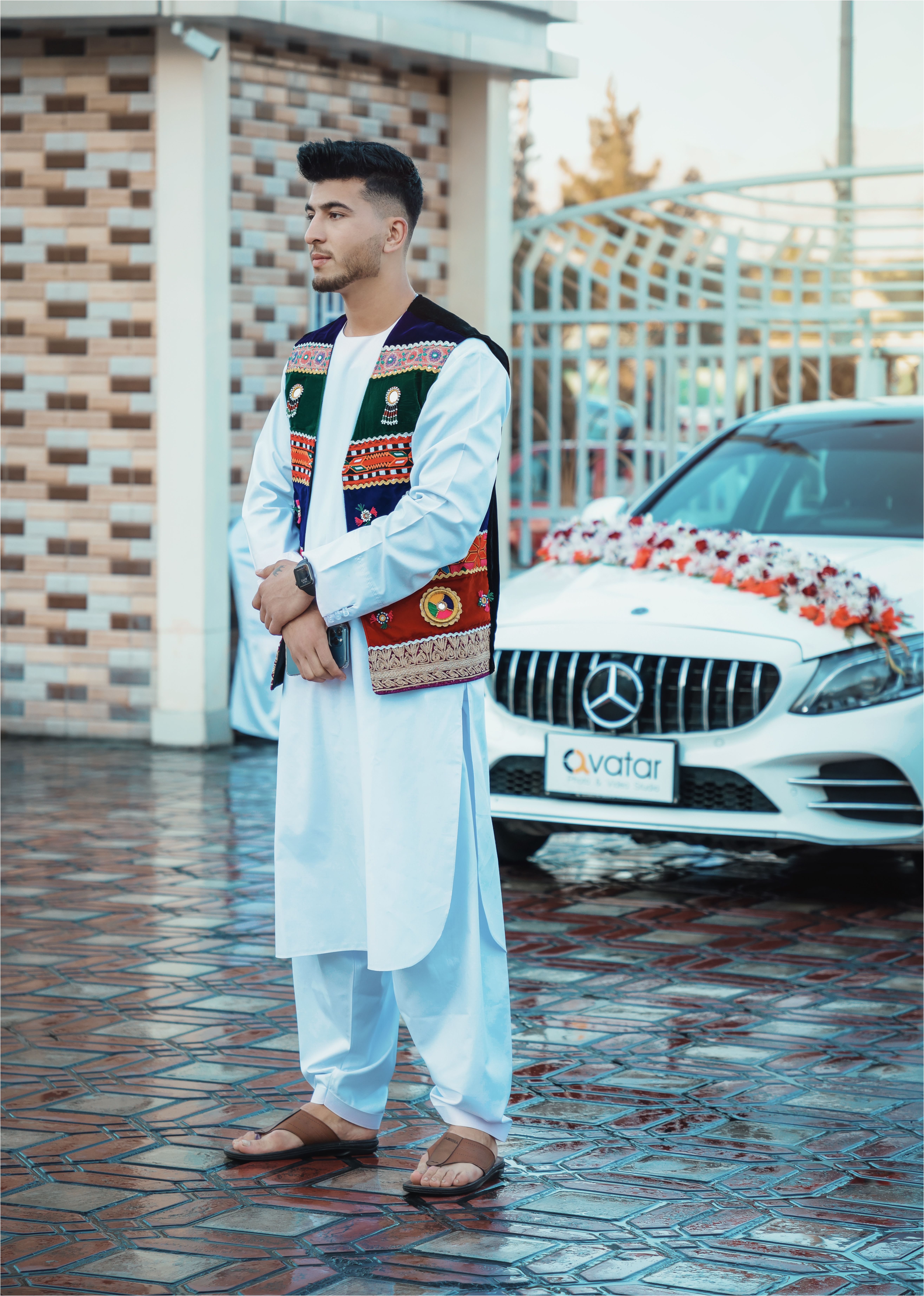 Man in traditional attire stands by a decorated car at a festive event, outdoors.