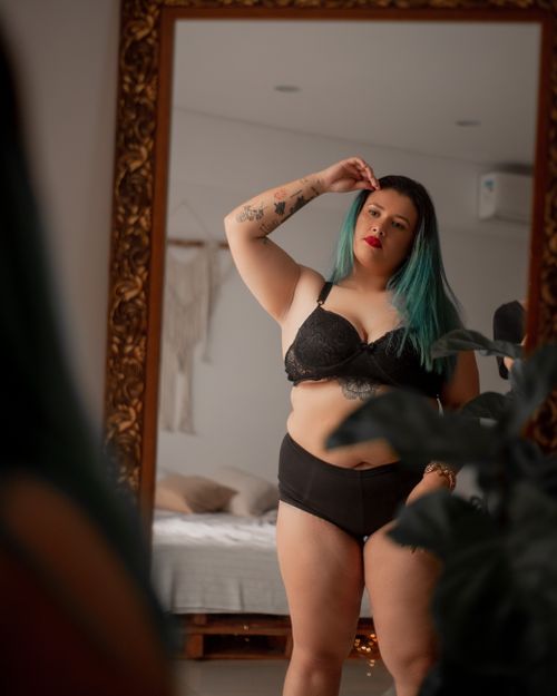 Free Plus size woman with blue hair standing confidently in front of a mirror, expressing body positivity. Stock Photo