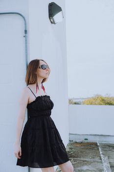 Elegant young woman posing in black dress and sunglasses against a modern outdoor backdrop.