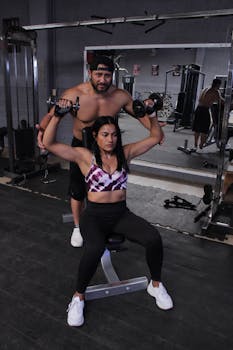 Man and woman performing shoulder workout with dumbbells in a gym setting.